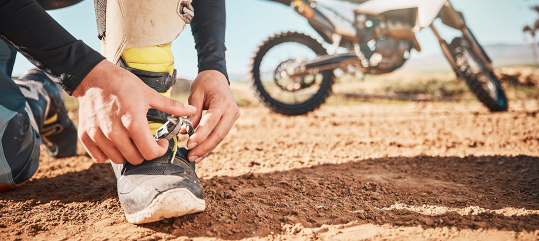 Person crouched tying motocross boots with motorbike in background.