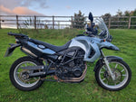 A silver BMW adventure motorcycle with black seat and tires parked on a green grassy field with a wooden fence and trees in the background.