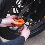 An orange ARMA lock on a motorcycle wheel being secured with a key by a person's hands, shown in a real-world outdoor setting.