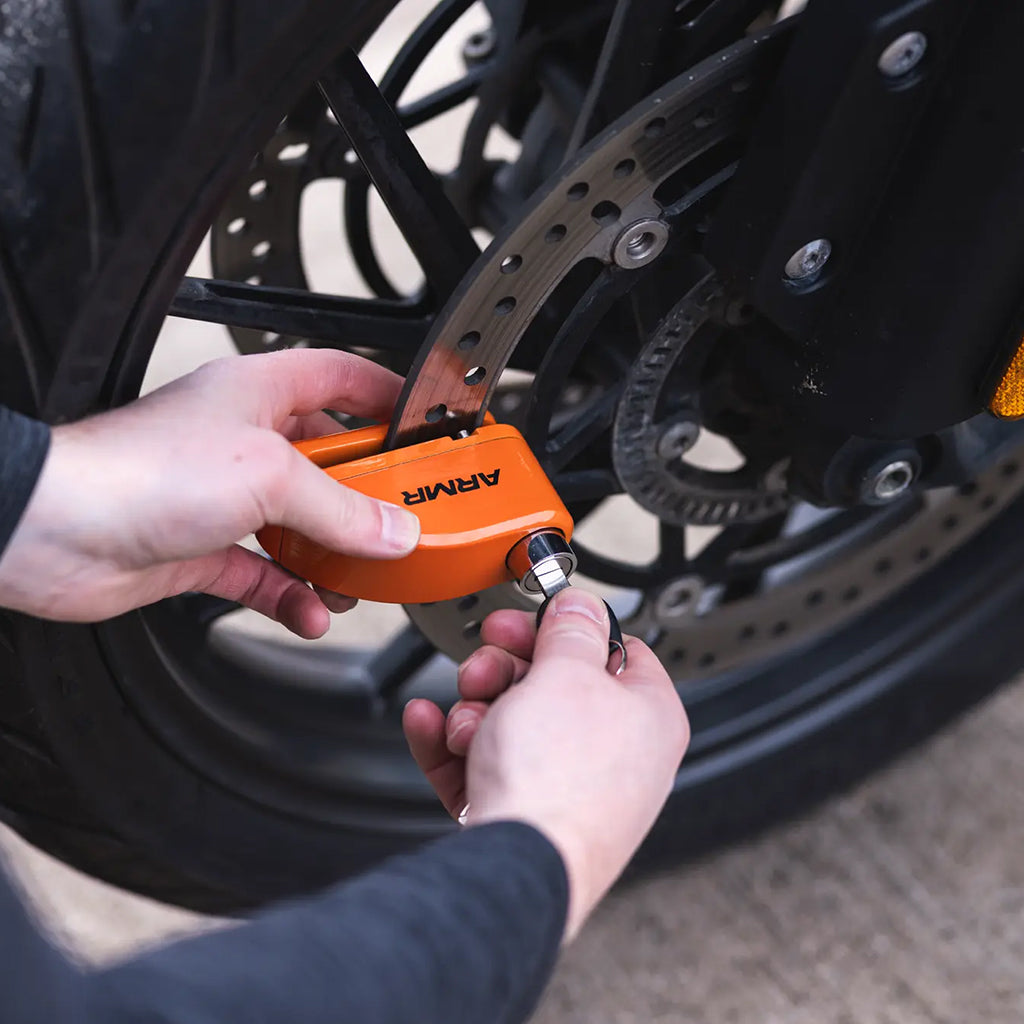 An orange ARMA lock on a motorcycle wheel being secured with a key by a person's hands, shown in a real-world outdoor setting.