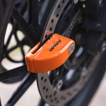 An orange ARMI smart lock with black branding is shown attached to a bicycle disc brake assembly in a close-up shot.