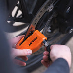 An orange ARMIA motorcycle lock is being secured with a black-handled tool by a person's hands near a motorcycle's wheel and brake disc.