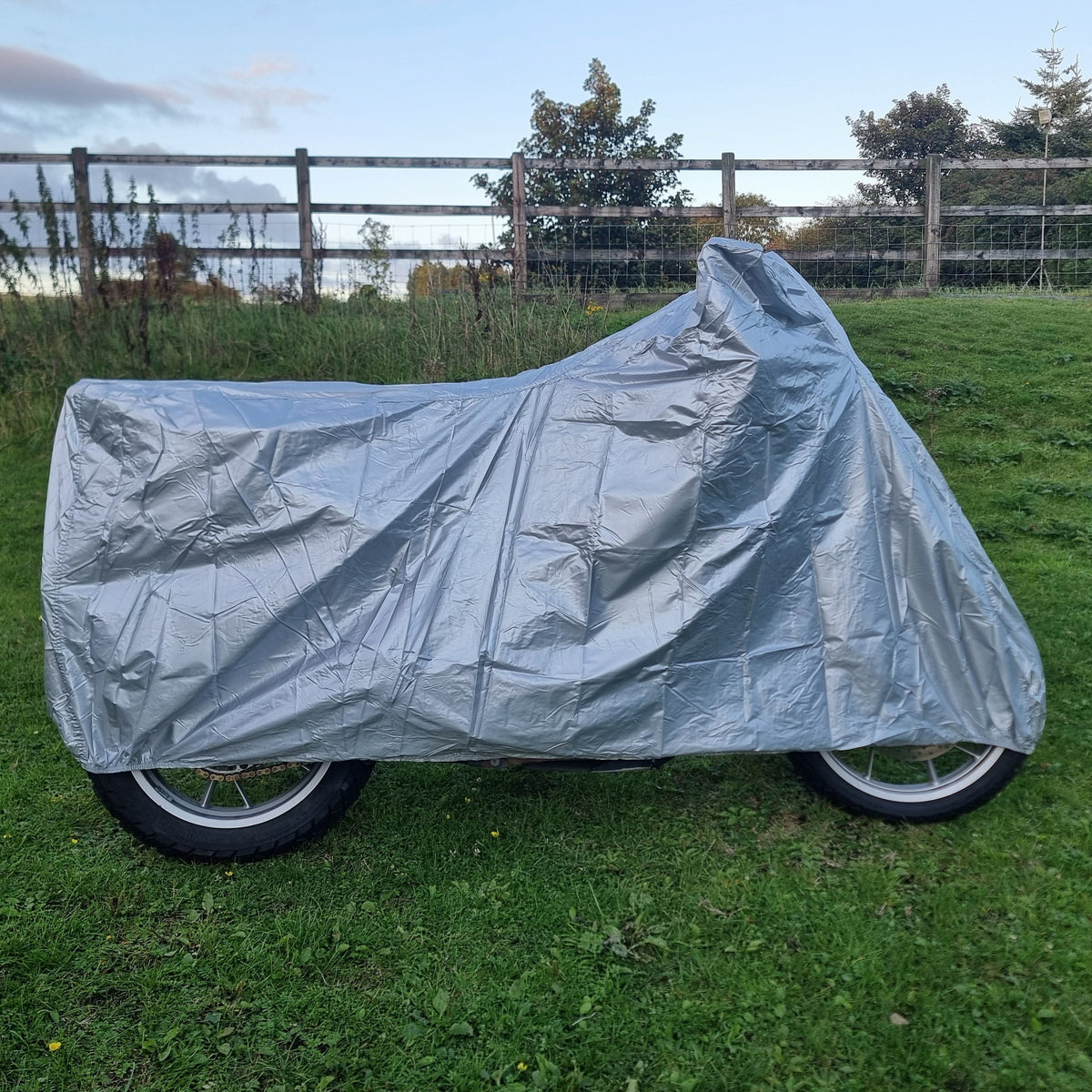 A silver motorcycle cover is draped over a motorcycle on a green grassy field, with a wooden fence and trees in the background under a blue sky.